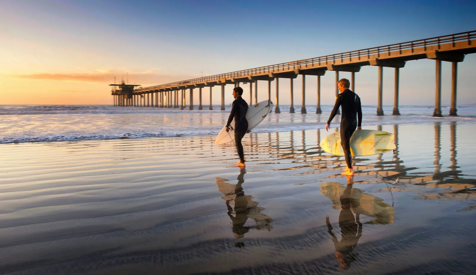 Two surfers carrying boards across the wet sand at Scripps Pier in the first light of the morning