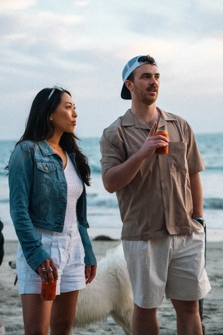 A couple standing at the water's edge at dusk with cans in hand, a white dog behind them and the ocean gone gray-blue