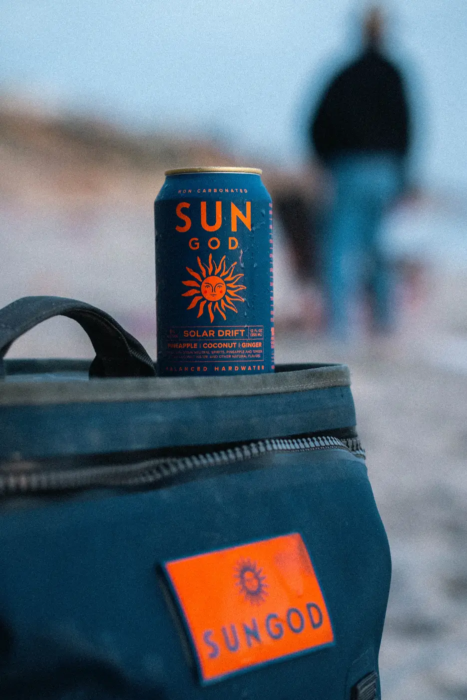 A can of Solar Drift standing in the top of a navy SUNGOD cooler bag on the sand, someone walking toward the water behind it