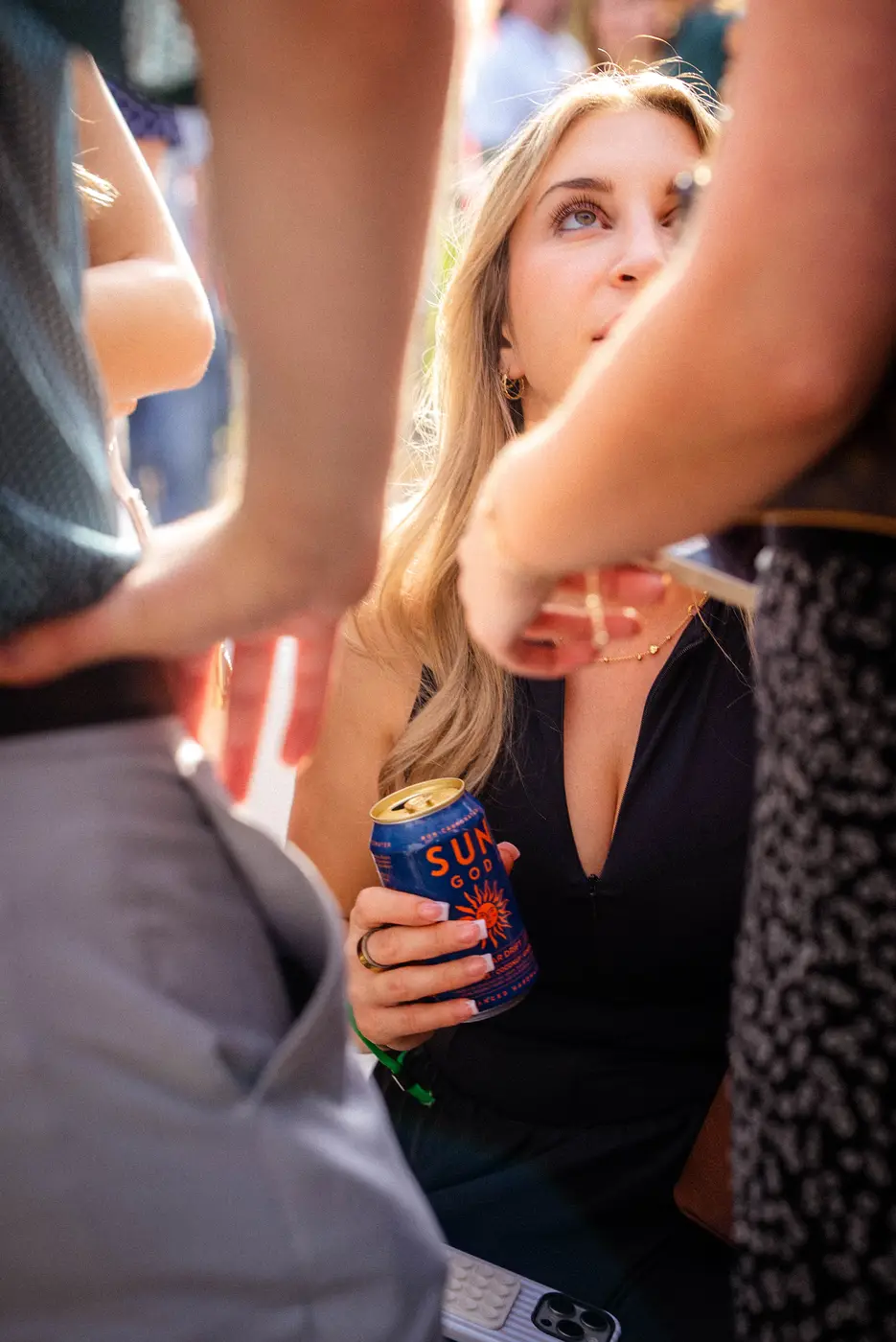 A woman in a black dress holding a can of Solar Drift in a crowd at a party, friends leaning in around her