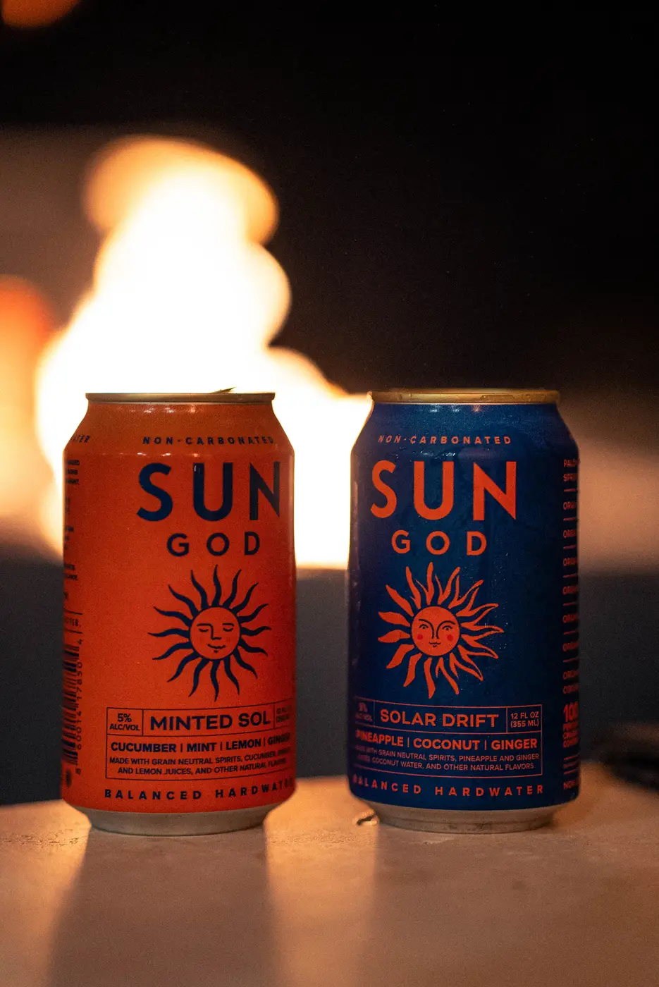 A Minted Sol and a Solar Drift on a table in front of a fire, the flames out of focus behind the cans