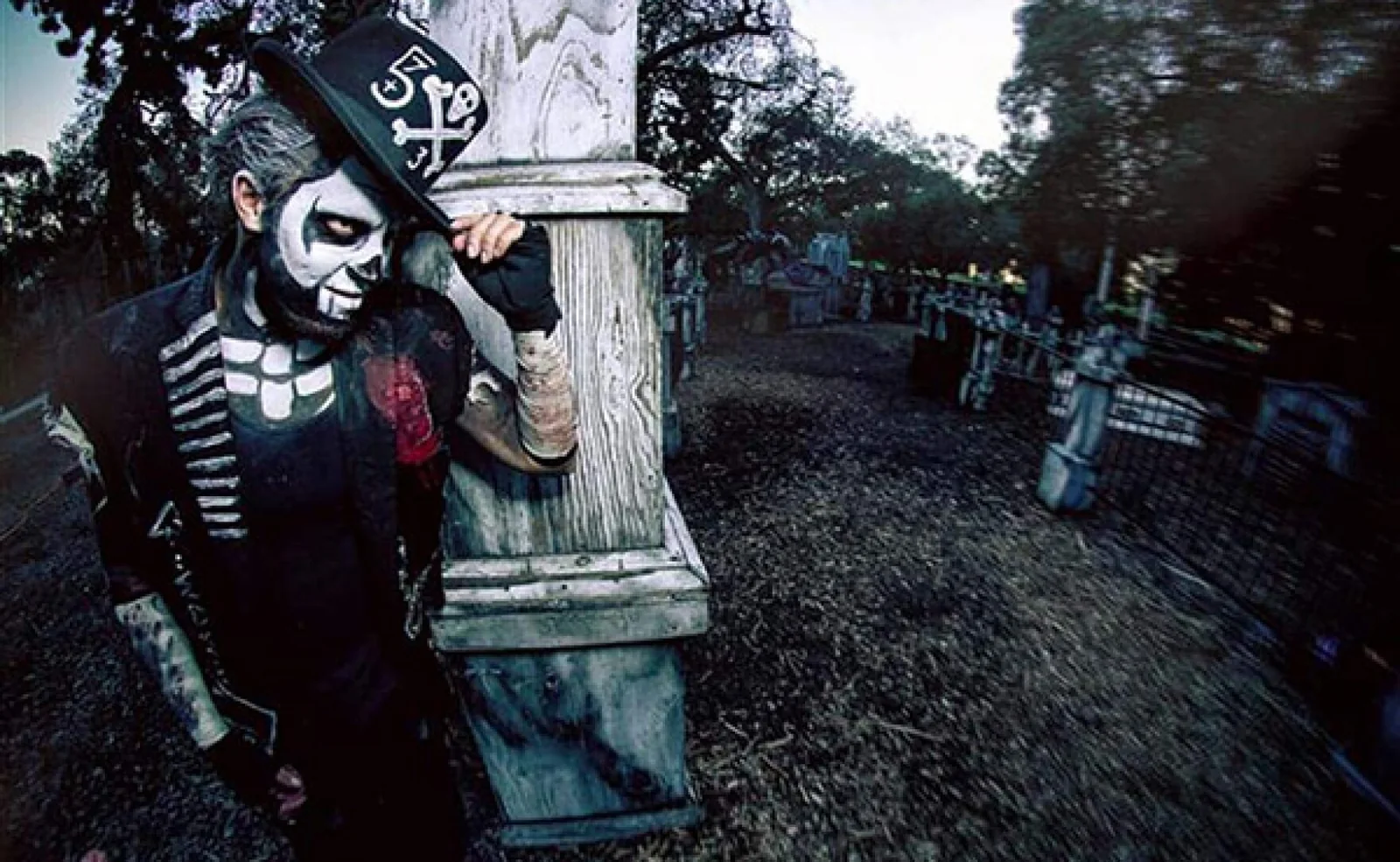 A skeleton-painted actor in a top hat leaning out from a tree on the Haunted Trail of Balboa Park at dusk