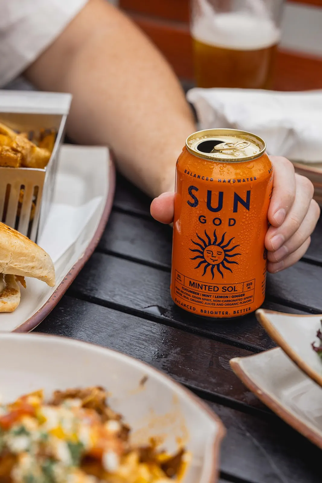 An orange can of SUNGOD Minted Sol on a dark wood table at lunch, beside a steak sandwich, fries and a bowl of loaded nachos