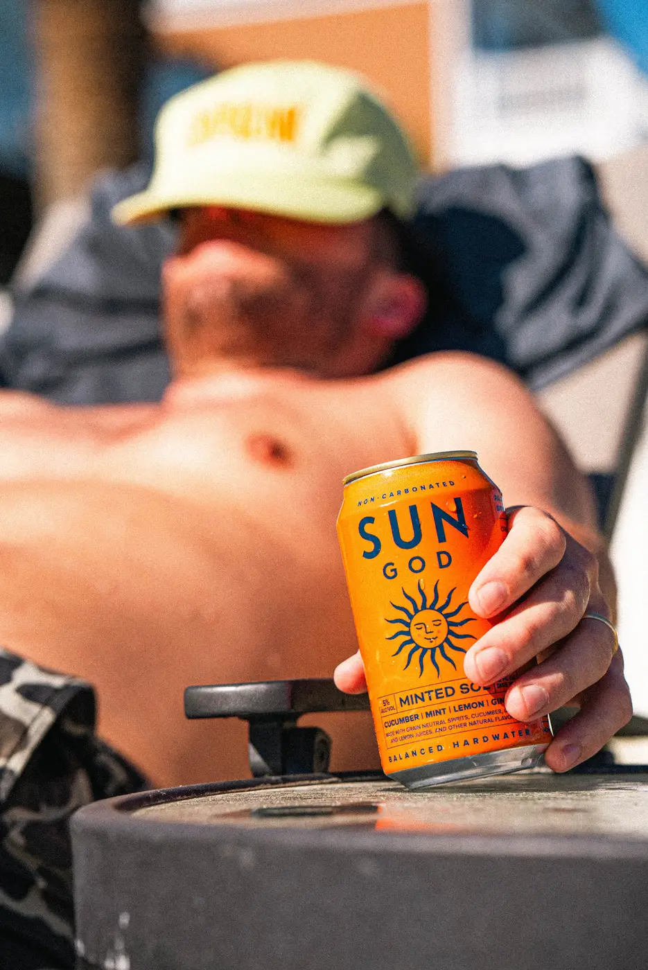 A guy in a yellow cap lying back on a lounge chair in full sun, a can of SUNGOD Minted Sol in his hand