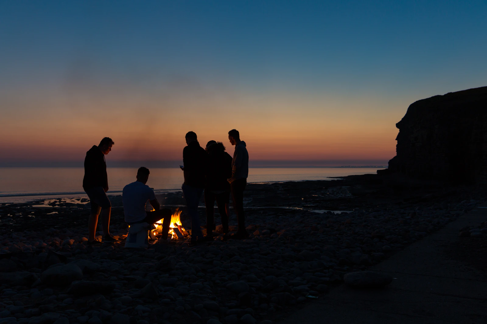 Five friends silhouetted around a beach fire after sunset, the sky still orange over the water