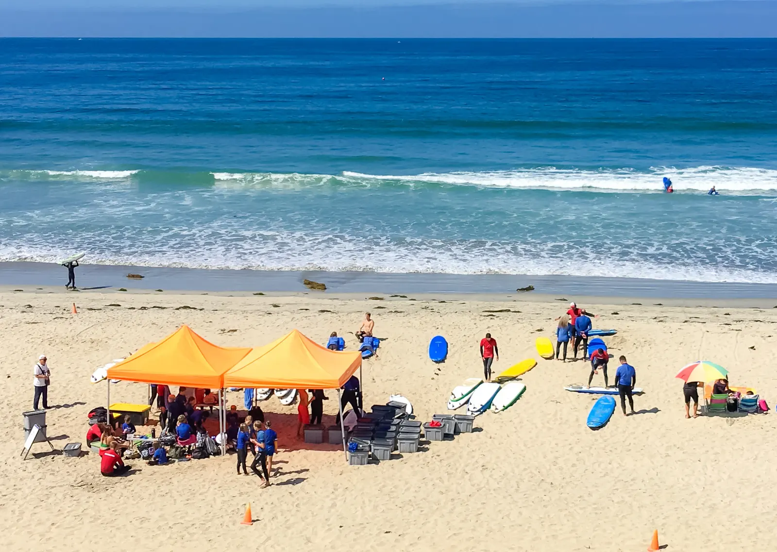 A surf school set up on the sand under two orange canopies, soft-top boards lined up and a group in wetsuits heading for small waves
