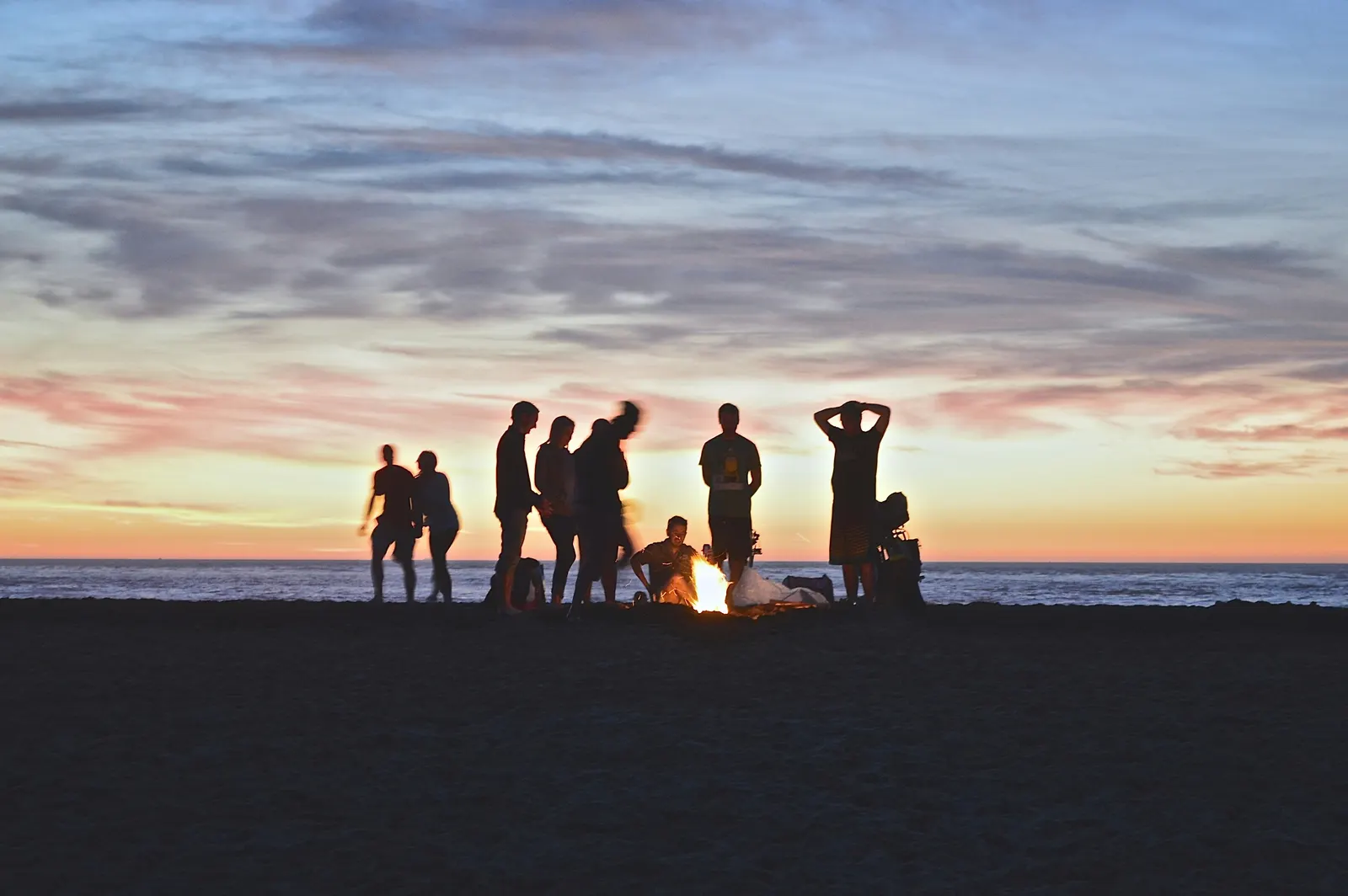 A group gathered around a beach bonfire at dusk with the sky going orange over the water