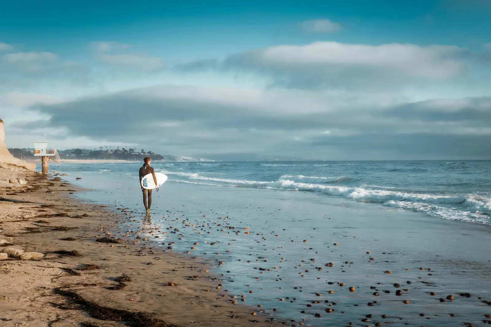 A surfer walking the wet sand at an Encinitas beach, a lifeguard tower and the bluffs behind